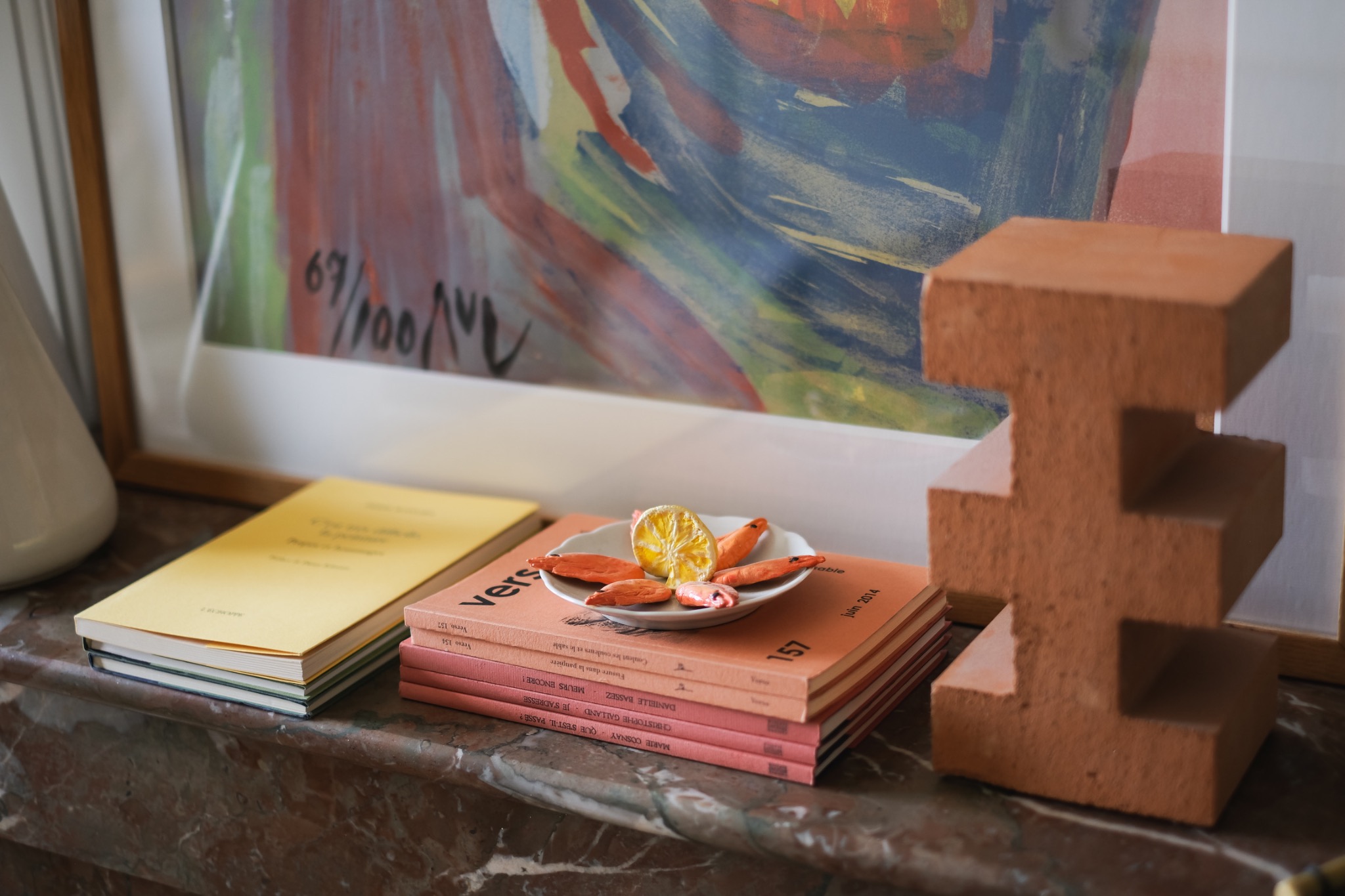 A curated material vignette with books, a small plate of sculptural objects, a framed artwork and a terracotta architectural form.