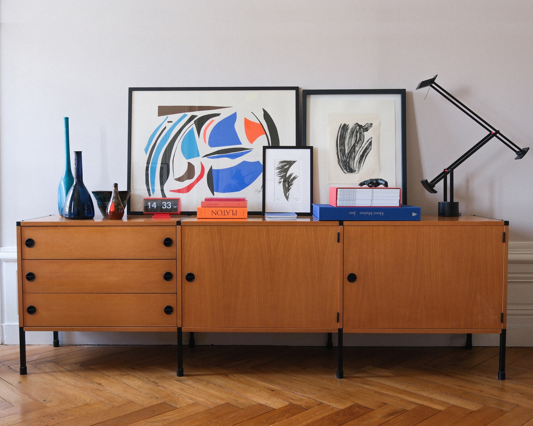 A refined interior vignette with a wooden sideboard, art, books, sculptural objects and a black task lamp.